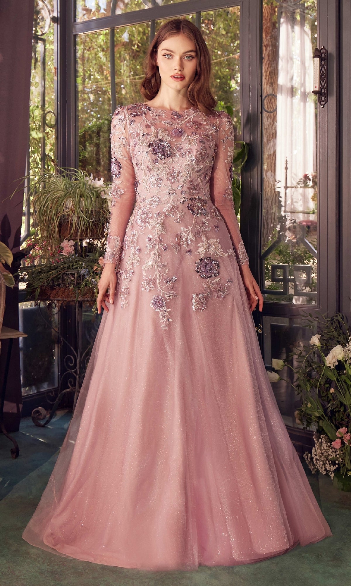 A woman stands indoors near large windows and plants wearing the Long Formal Pink Dress: Andrea and Leo A1356, featuring an A-line skirt with pink fabric adorned with floral sequin embroidery.